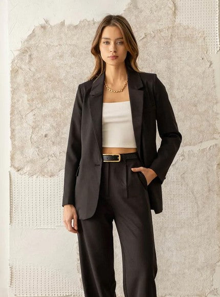 Classic black tailored single-breasted blazer  with notched lapel and pocket with a flap. Brunette model is wearing it with a white top and classic black trousers.