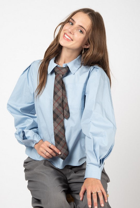 Sky blue collared button-down shirt with puff sleeves. The model is wearing it with gray trousers, a gray and red plaid tie, and silver jewelry. She is sitting in front of a white background.