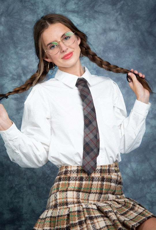 Off-white collared button-down shirt with puff sleeves. The model is wearing it with a brown, beige, and green tweed skirt and a gray and red plaid tie. She has her hair in two loose braids, and is posing for a mock school photo in front of a blue backdrop.