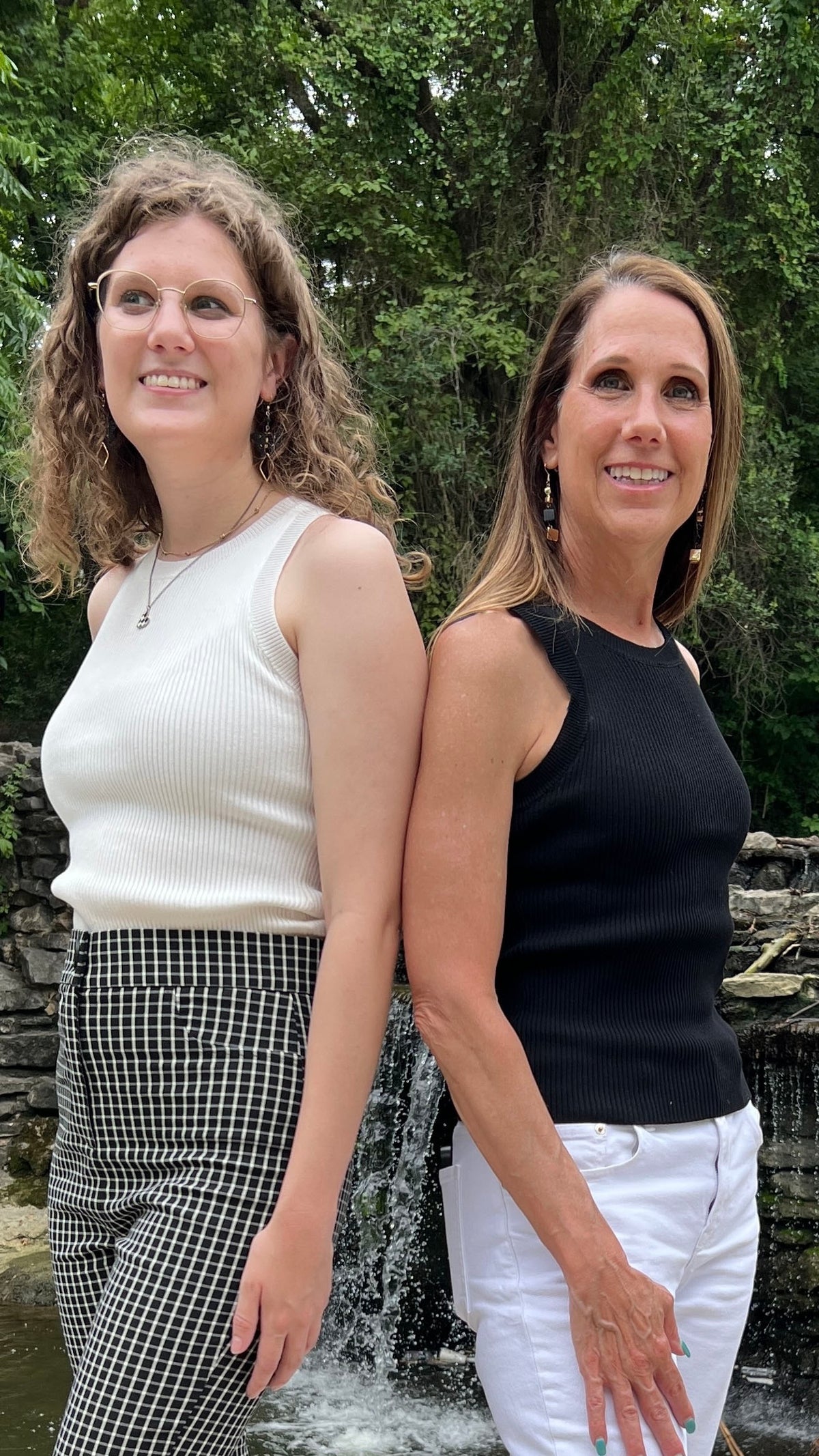 Racer-back style tank top with a crew neck in white or black colors. Two models: one is wearing white racerback tank with black checked pants and the other is wearing the black racerback tank with white denim. Models are standing in a wooded area facing the camera.