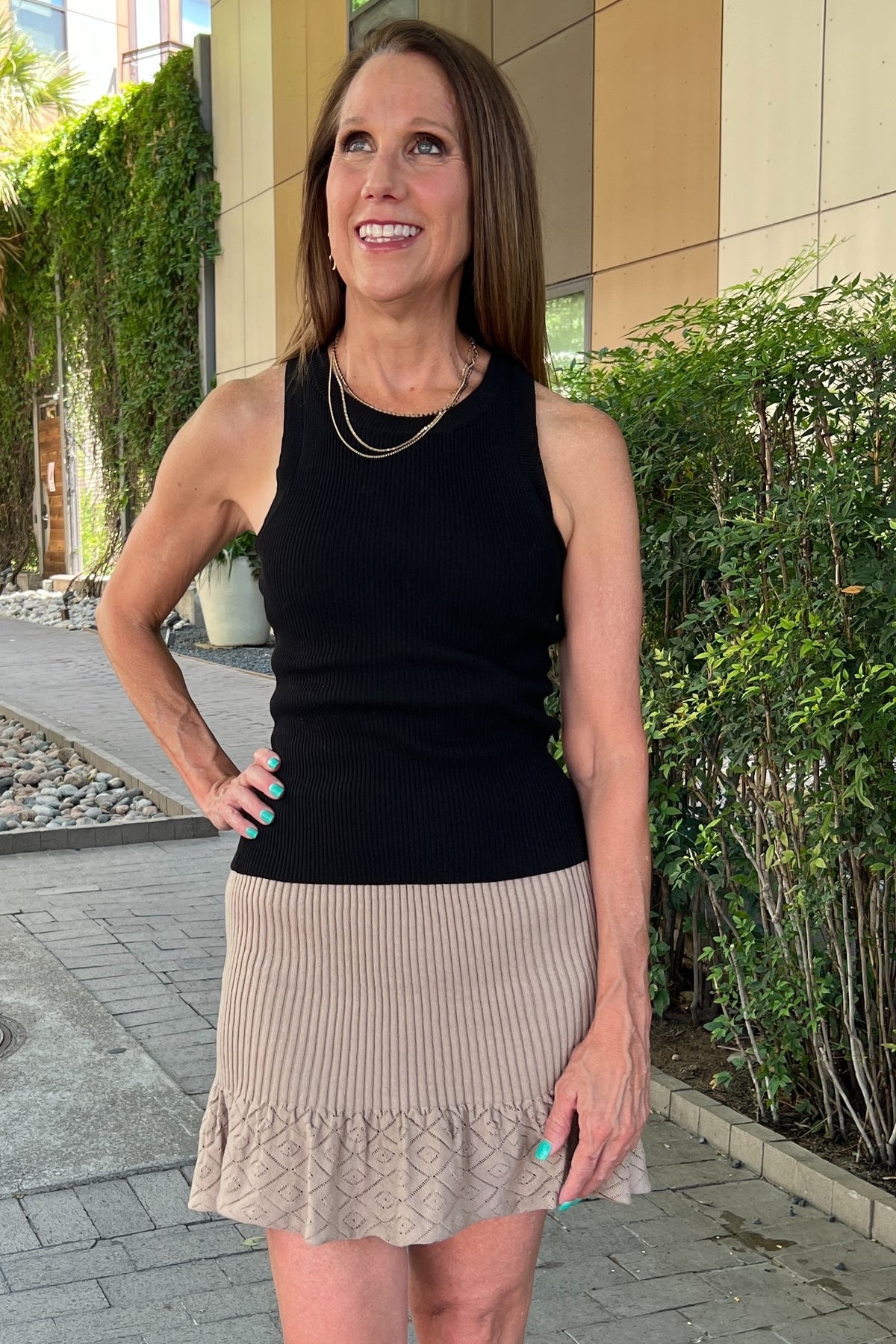 Taupe knit mini skirt with ribbed texture and ruffle 3 inch hem. Model is wearing it with a black tank top and multistrand necklace. She's standing on a sidewalk.