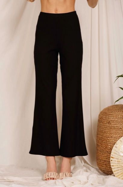 Model wearing black ribbed flare leg pants. The model is shown from the waist down standing in front of a white curtain wearing the pants with no shirt and with nude heels.