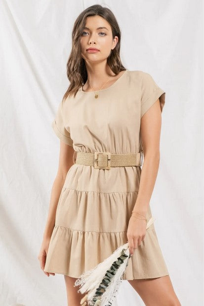 Taupe-colored tiered ruffle mini dress. Dress has cuffed cap sleeves and a sccoped neckline and back button closure. Comes with a straw belt. Model is standing in front of a white wall.