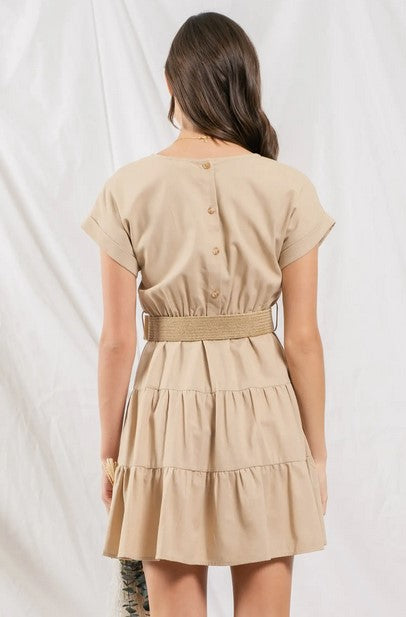Taupe-colored tiered ruffle mini dress. Dress has cuffed cap sleeves and a sccoped neckline and back button closure. Comes with a straw belt. Model is standing in front of a white wall.