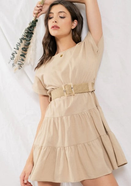 Taupe-colored tiered ruffle mini dress. Dress has cuffed cap sleeves and a sccoped neckline and back button closure. Comes with a straw belt. Model is standing in front of a white wall.