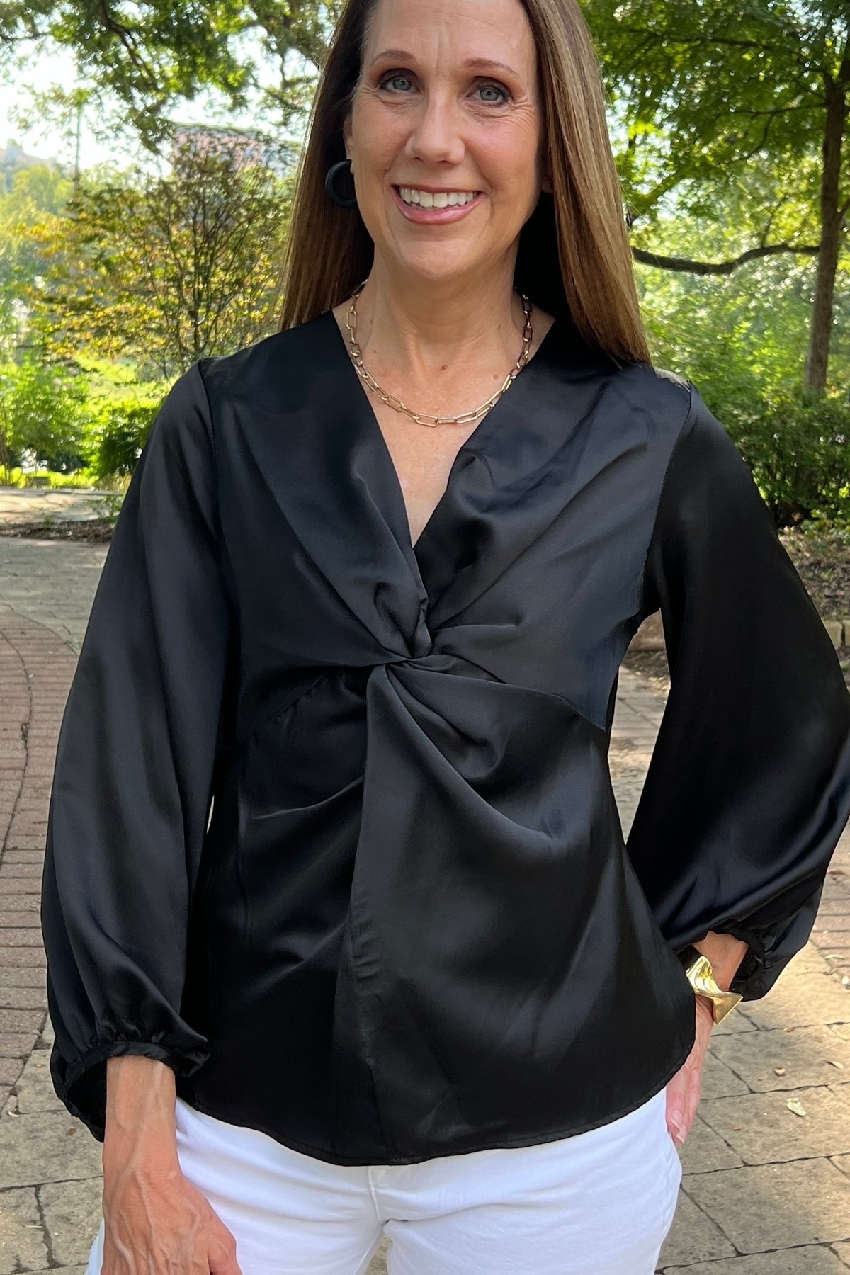 Black V-neck satin blouse with a twisted detail in the front. The model is wearing it with white jeans and sandals.