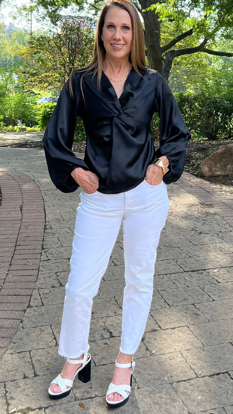 Black V-neck satin blouse with a twisted detail in the front. The model is wearing it with white jeans and sandals.