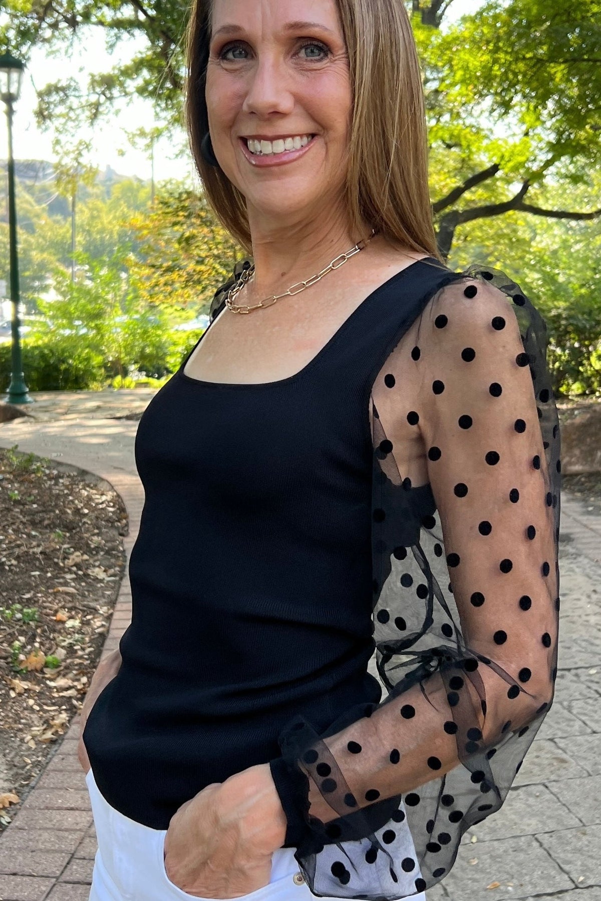 Dressy black fitted top with sheer sleeves that have a swiss-dot pattern. Model is wearing it with linen shorts. 