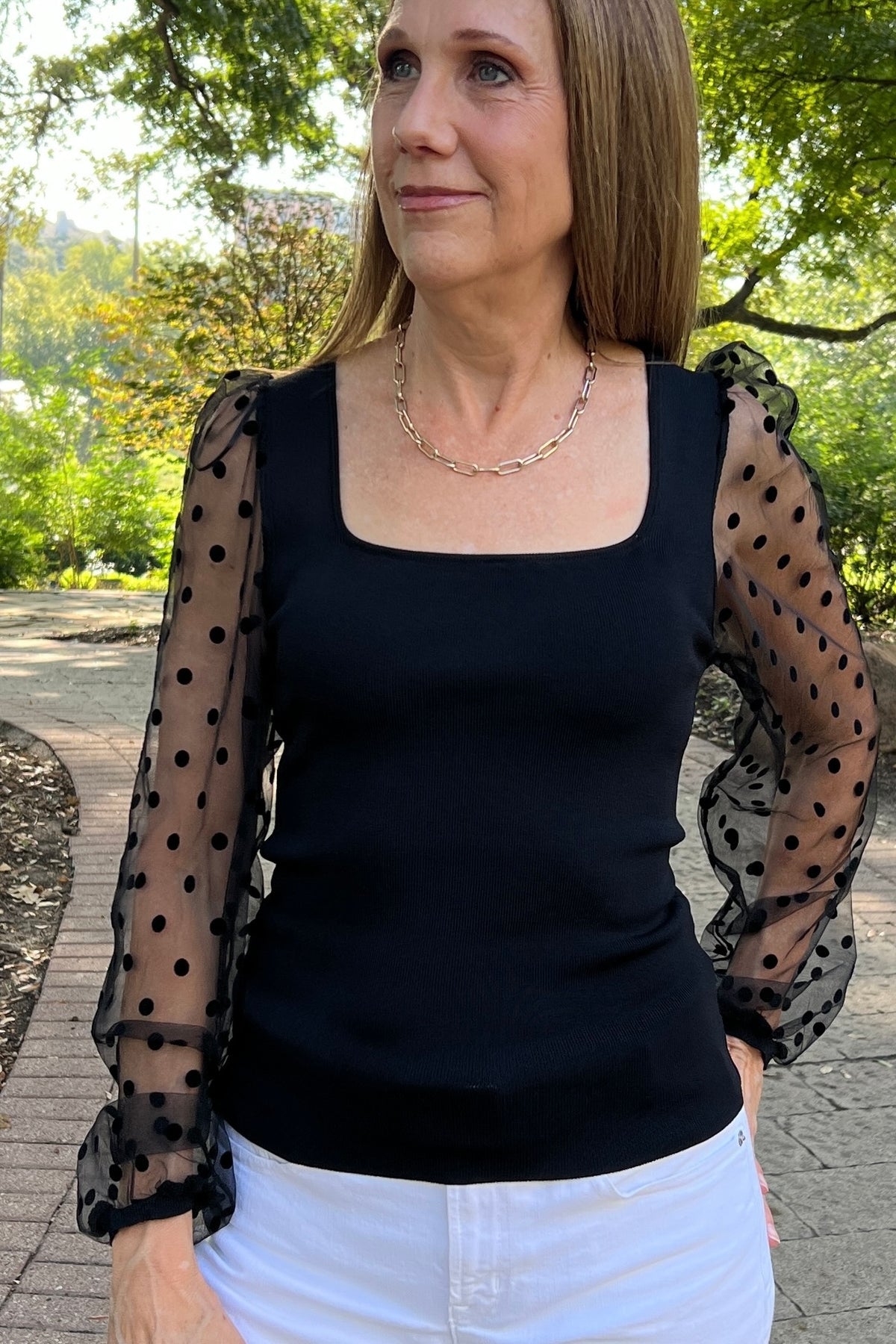 Dressy black fitted top with sheer sleeves that have a swiss-dot pattern. Model is wearing it with white jeans. 