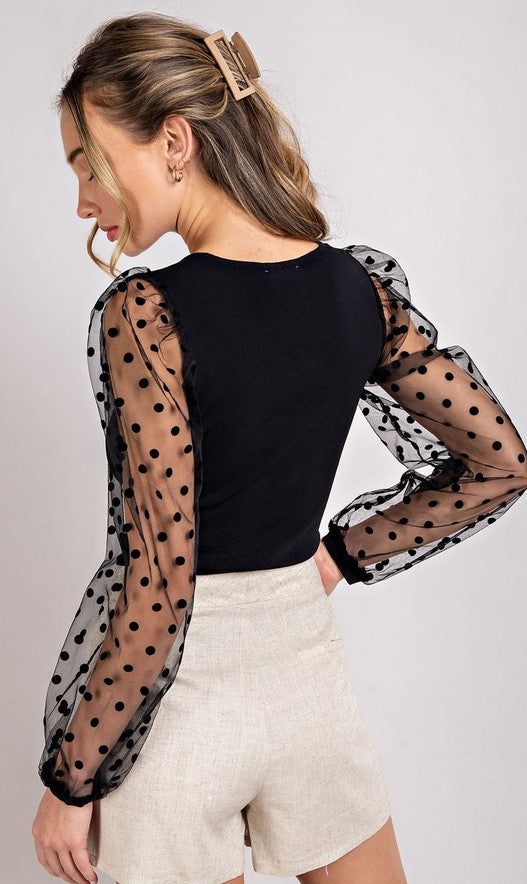 Dressy black fitted top with sheer sleeves that have a swiss-dot pattern. Model is wearing it with linen shorts. 