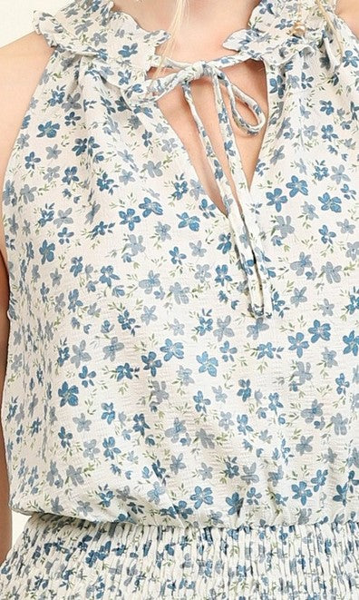 Close up of sleeveless top or blouse with a smocked waist and ruffle tie neckline has a denim blue ditsy floral pattern on an ivory or white base.