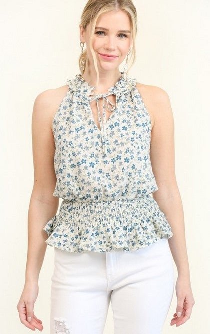 Sleeveless top or blouse with a smocked waist and ruffle tie neckline has a denim blue ditsy floral pattern on an ivory or white base. Model is blonde and wearing the top with white denim and is standing in front of a white wall.