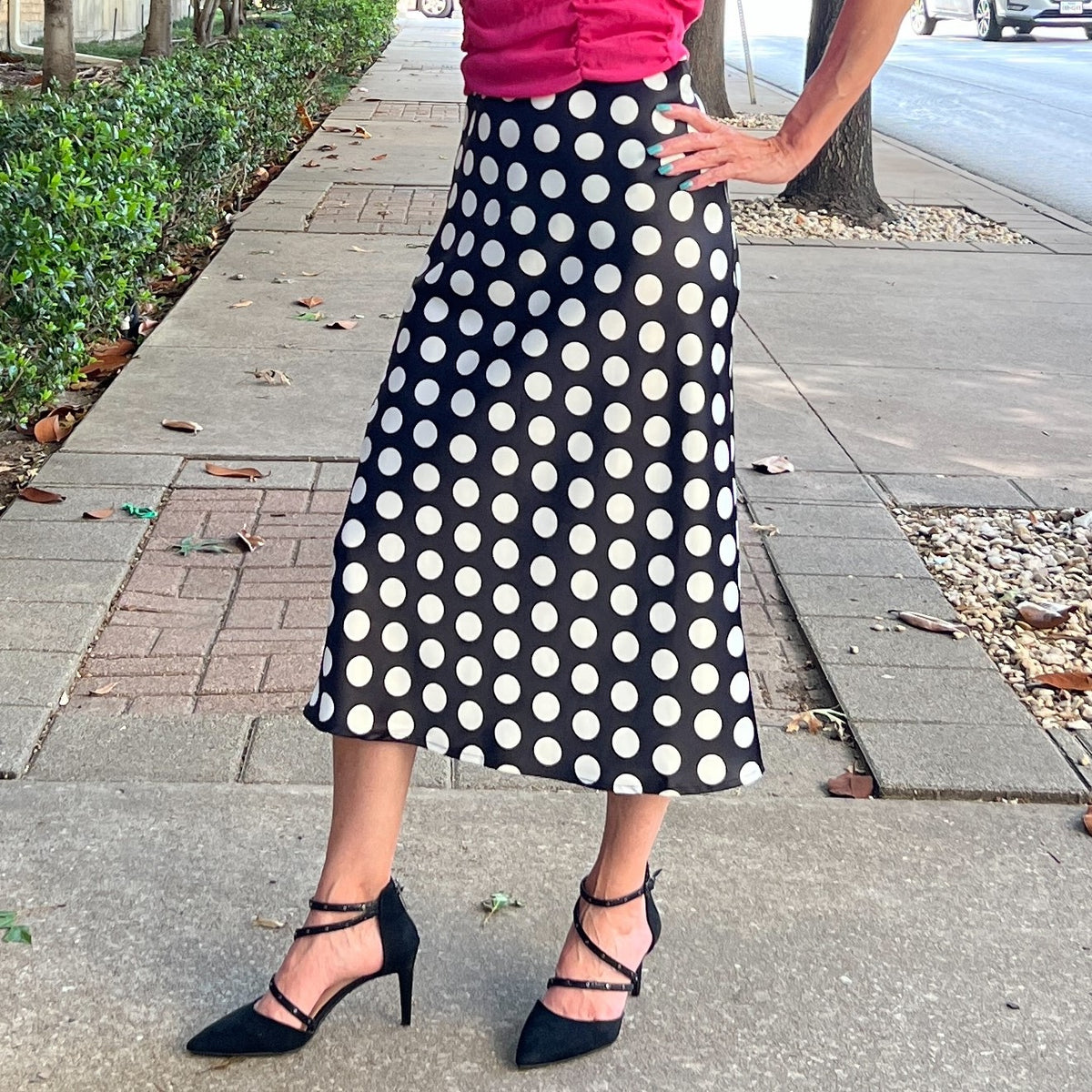 Midi length black satin skirt with medium white polka dots. Pencil skirt shape at hips with a slight a-line flare at the knees to calves. Model is wearing the black satin skirt with black heels and pink crop top and is standing on a sidewalk.