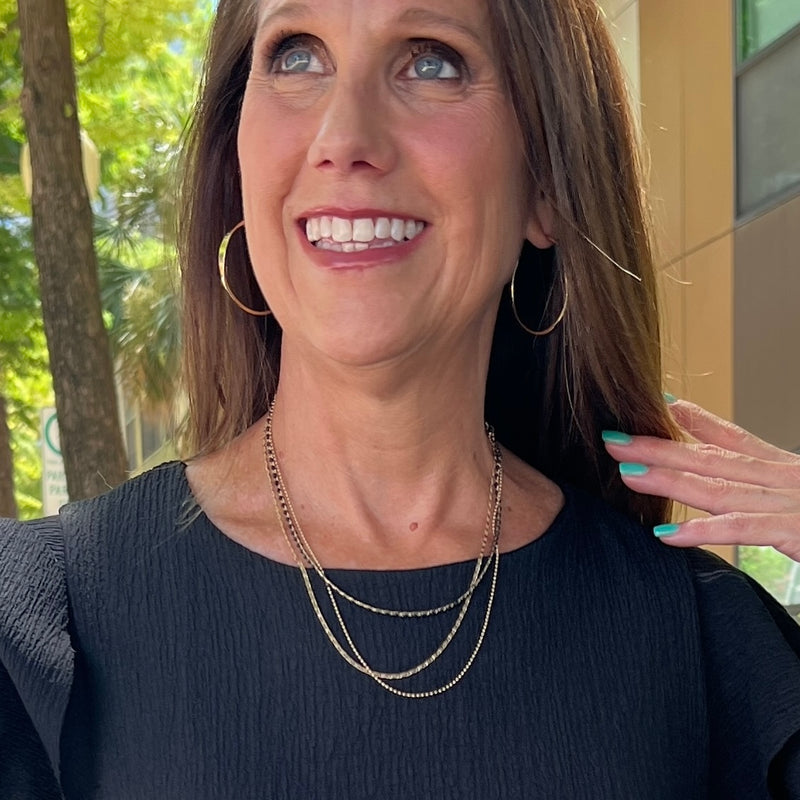 A brunette woman smiling wearing Gold hoop earrings and a triple-strand gold necklace with black beads. She's holding her hair back to show off the jewelry and is standing in front of some trees and a building. She is wearing a black top.