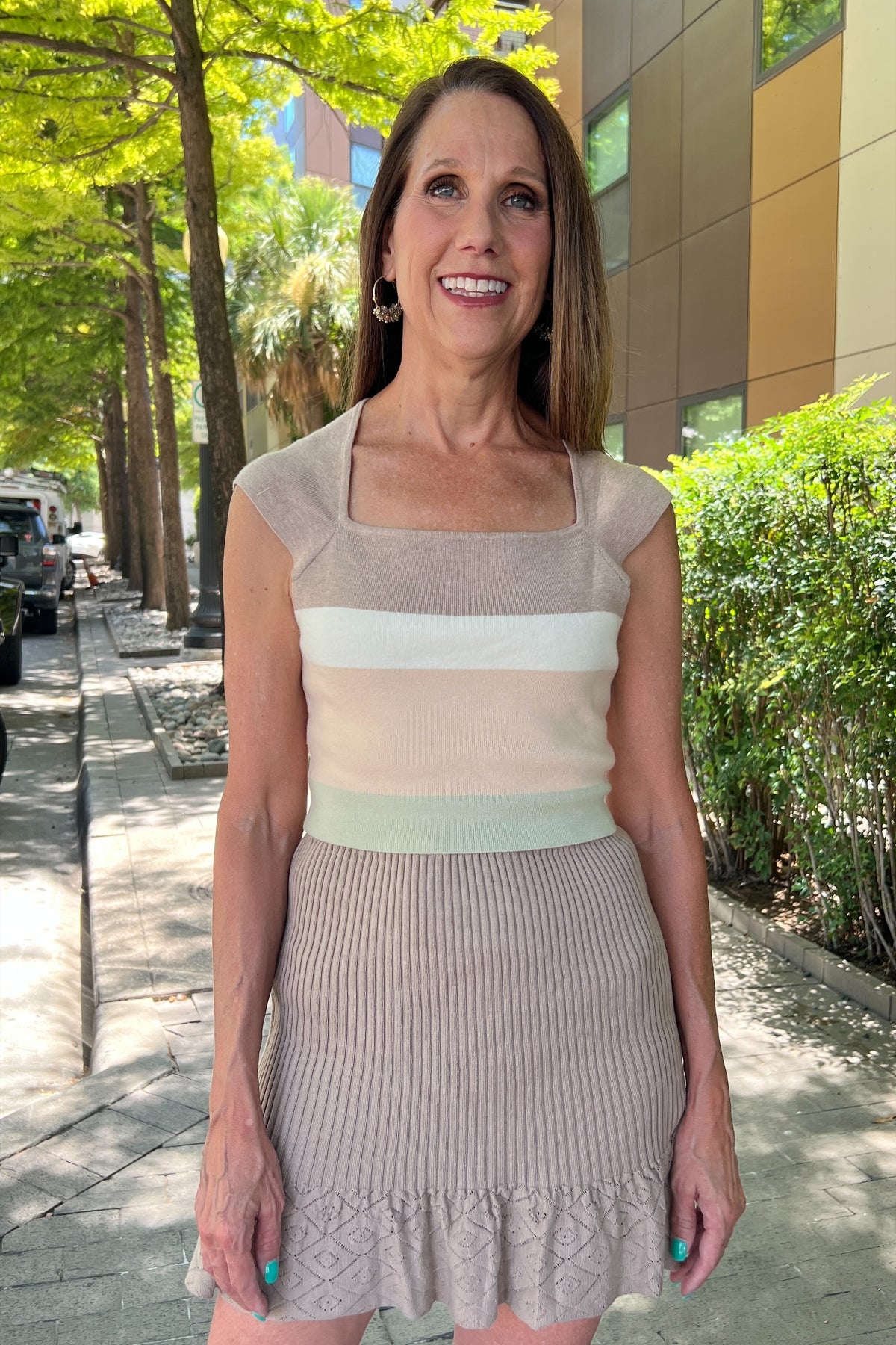 Taupe knit mini skirt with ribbed texture and ruffle 3 inch hem. Model is wearing it with a cropped shirt with taupe, butter, sage, and beige stripes. She's standing on a sidewalk. 