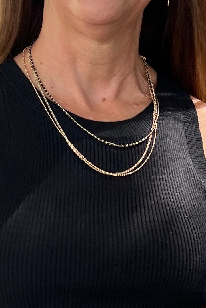 A multi-strand necklace of two gold chains and one black-beaded strand. Pictured on a close up of a woman wearing a black top.