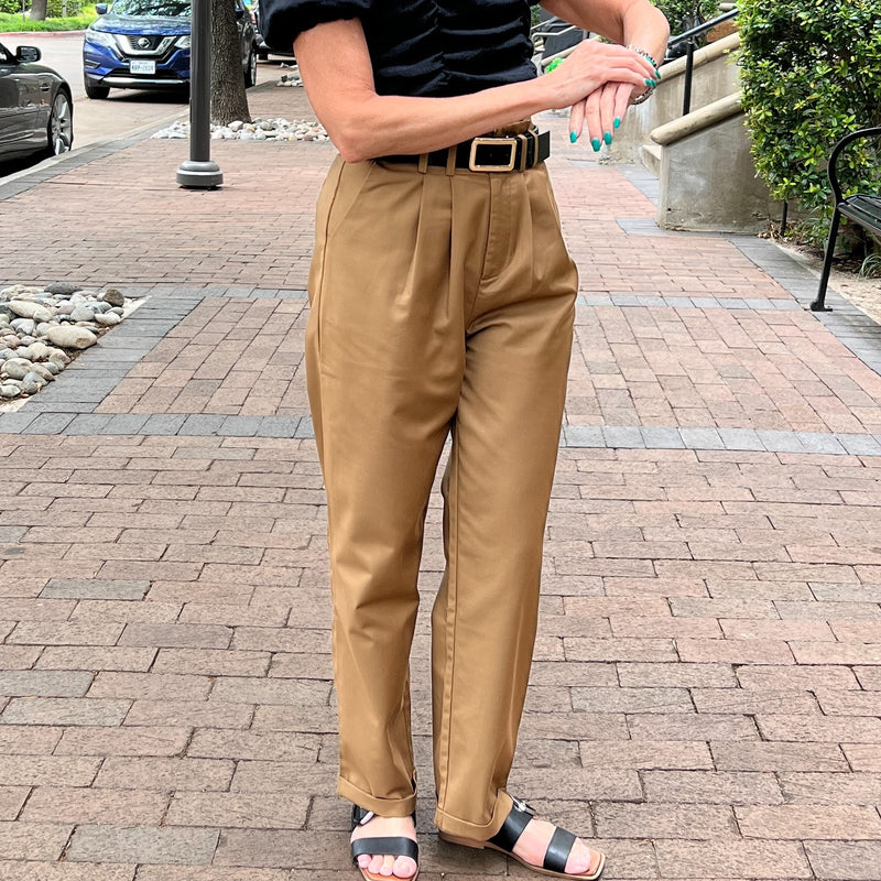 Caramel colored pleated high waist pants have a cuffed hem and side pockets. Model is wearing the pants with a black crop top, belt and sandals and is standing on a sidewalk.