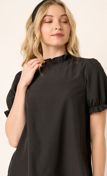 Black short sleeved mock neck shirt with a ruffle detail at the neck and sleeves. The model is wearing it with a black headband. She is posing in front of a cream wall.