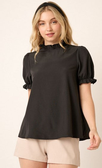 Black short sleeved mock neck shirt with a ruffle detail at the neck and sleeves. The model is wearing it with khaki shorts and a black headband. She is posing in front of a cream wall.