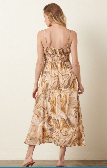 Olive, taupe, and beige marble patterned sundress has a ruched empire waist with zipper in back and adjustable straps. The dress is midi length. Model is wearing it in front of a beige wall with tan sandals and gold hoop earrings. 