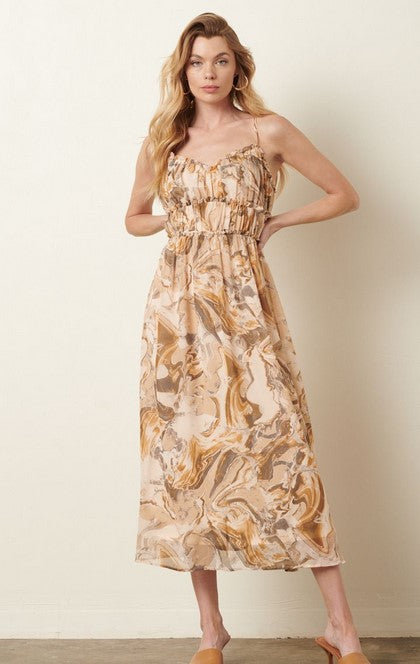 Olive, taupe, and beige marble patterned sundress has a ruched empire waist with v-style neckline and adjustable straps. The dress is midi length. Model is wearing it in front of a beige wall with tan sandals and gold hoop earrings. 