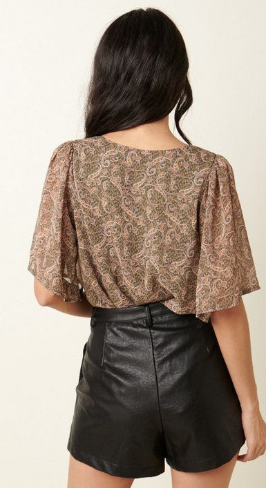 Olive green paisley v-neck blouse with loose flutter sleeves. The model is wearing it with black leather shorts and posing facing away from the camera in front of a cream wall.