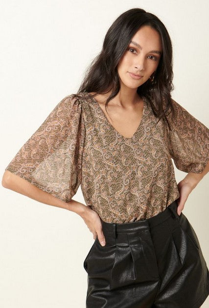 Olive green paisley v-neck blouse with loose flutter sleeves. Lined bodice with a cream and caramel paisley pattern on an olive background.The model is wearing it with black leather shorts and posing in front of a cream wall.