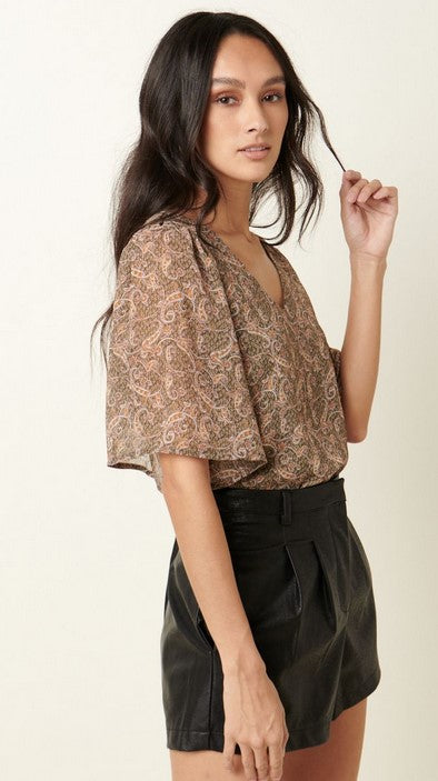 Olive green paisley v-neck blouse with loose flutter sleeves. The model is wearing it with black leather shorts and posing in front of a cream wall.