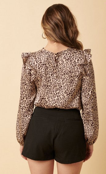 Long sleeved beige and brown leopard print shirt with a mock neck and ruffle shoulders. The model is wearing it with black shorts and posing facing away from the camera in front of a cream background.