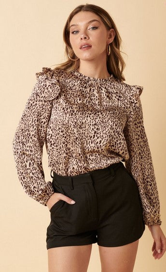 Long sleeved beige and brown leopard print shirt with a mock neck and ruffle shoulders. The model is wearing it with black shorts and posing in front of a cream background.