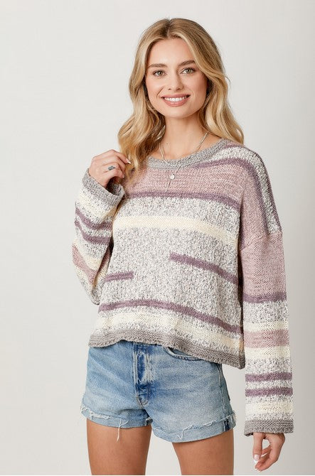 Cropped, striped spring sweater in lavender, gray, and ivory. Model is blonde, and is wearing the sweater with light-wash denim jean shorts, thin layered gold necklaces, and is standing in front of a white backdrop.
