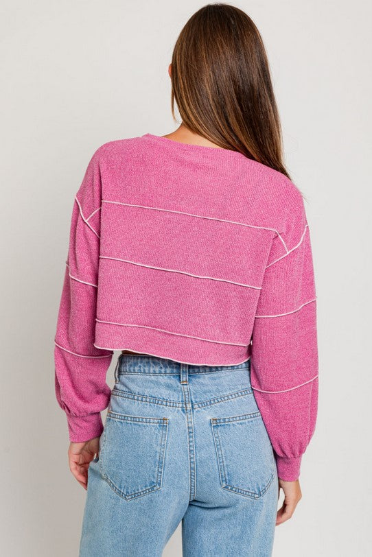 Back of pink cropped sweater textured with a ribbed knit fabric and lettuce hem banding in a lighter pink. Fit is cropped. Model is wearing it with button-fly jeans and is standing in front of a white background.