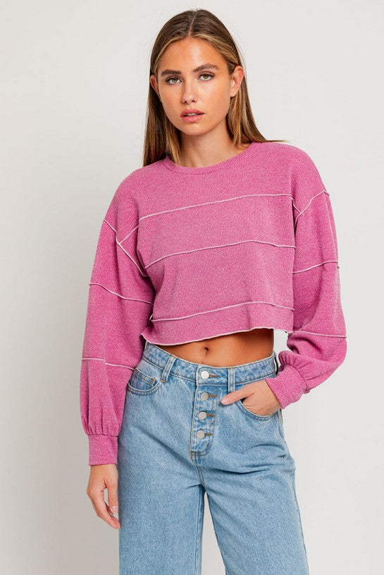 Pink cropped sweater textured with a ribbed knit fabric and lettuce hem banding in a lighter pink. Fit is cropped. Model is wearing it with button-fly jeans and is standing in front of a white background.