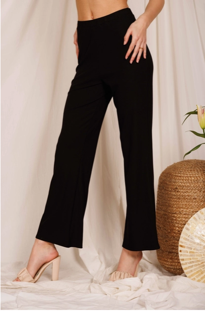 Model wearing black ribbed flare leg pants. The model is shown from the waist down standing in front of a white curtain wearing the pants with no shirt and with nude heels.