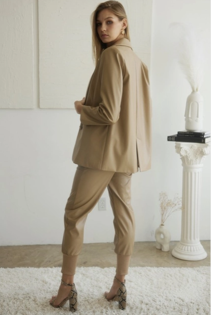 Camel oversize faux leather blazer worn with camel joggers and snakeskin high-heeled shoes. The model is facing away from the camera and looking back. She is in a room with white paneled walls and is standing on a fuzzy white rug.