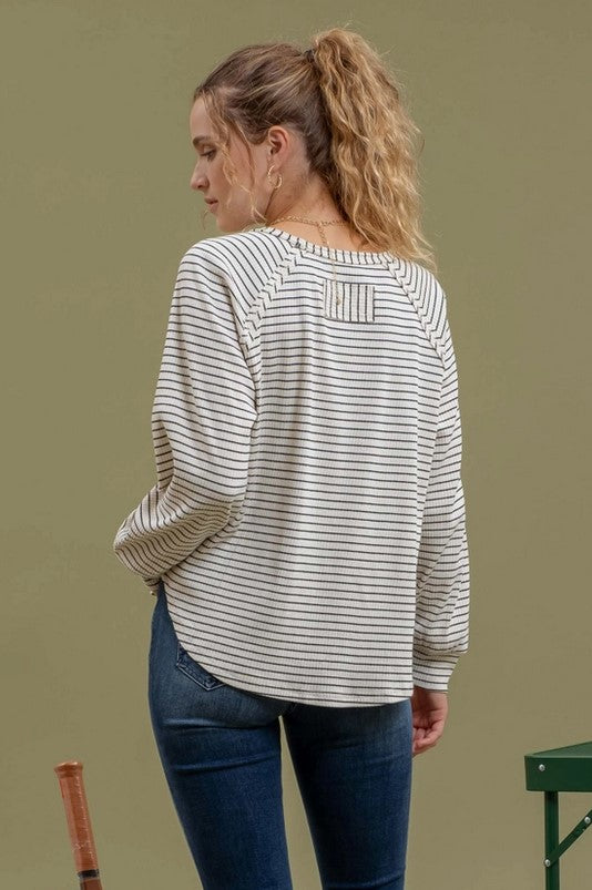 Back view of ivory raglan-style t-shirt with black stripes is being modeled by a blonde woman with a pony tail wearing jeans. She is standing near pickleball equipment.