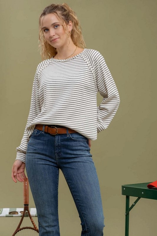 Ivory raglan-style t-shirt with black stripes is being modeled by a blonde woman with a pony tail wearing jeans. She is standing near pickleball equipment.