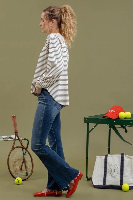 Side view of ivory raglan-style t-shirt with black stripes is being modeled by a blonde woman with a pony tail wearing jeans. She is standing near pickleball equipment.