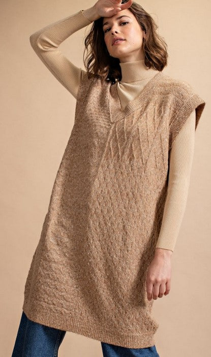 A camel colored sleeveless knit tunic dress with a V-neck. The model is wearing it over a camel turtleneck and blue denim jeans. She is posing in front of a tan background.