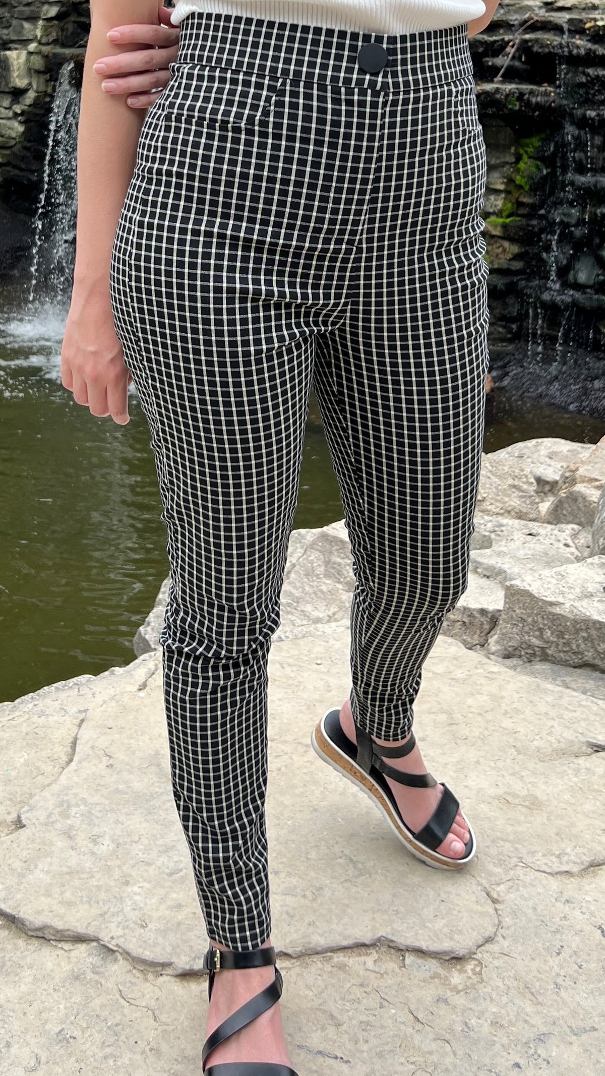 A woman modeling black and white checked skinny high-waisted pants in front of a waterfall. We see her from the waist down, and she has tucked a white shirt into the pants and is wearing black sandals.