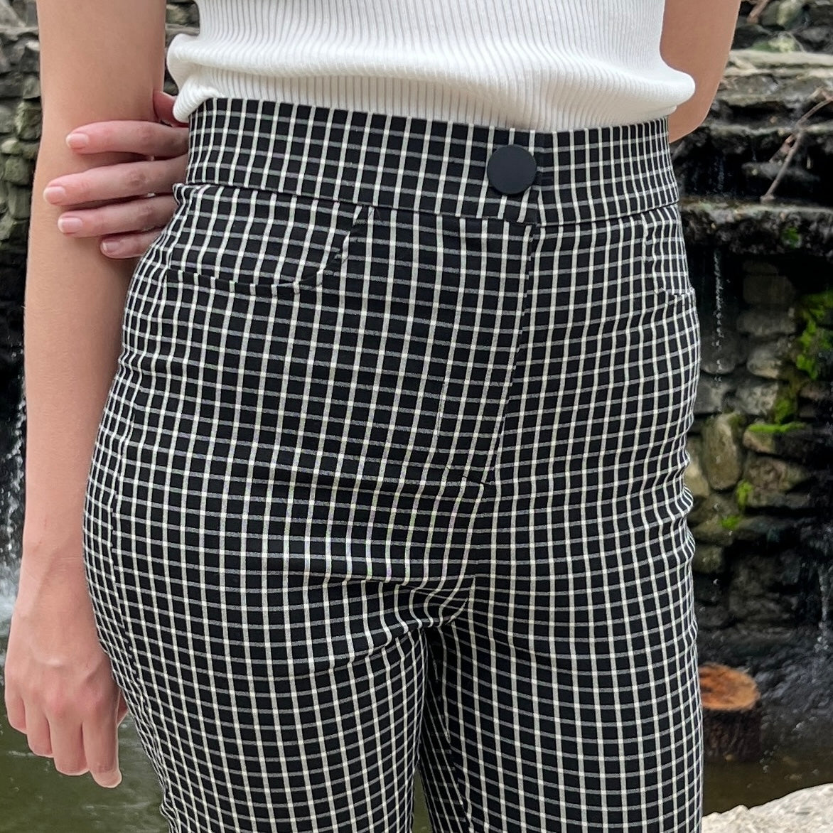 A woman modeling black and white checked skinny high-waisted pants in front of a waterfall. We see her from the waist to the knees, and she has tucked a white shirt into the pants.