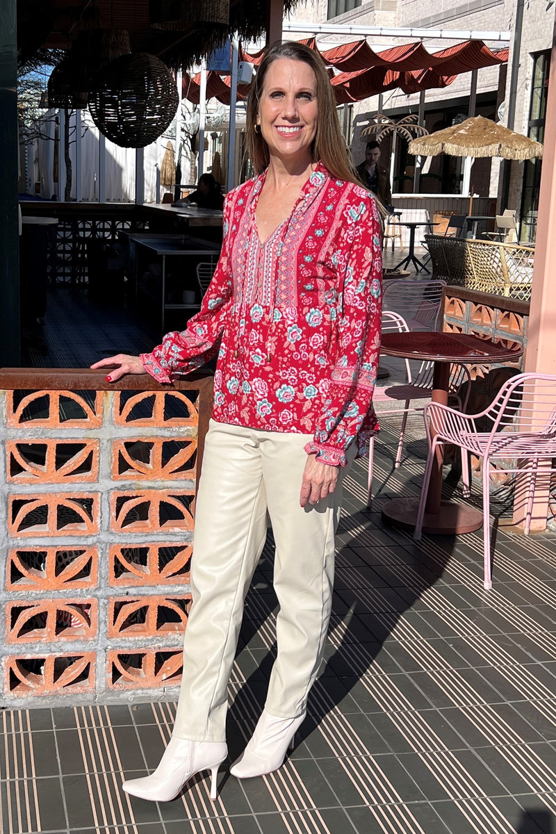 Red blouse with blue and white floral patterned blouse with long sleeves. Has a babydoll cut and thin tie neckline. Model is wearing it with beige leather pants and is standing in front of a restaurant.