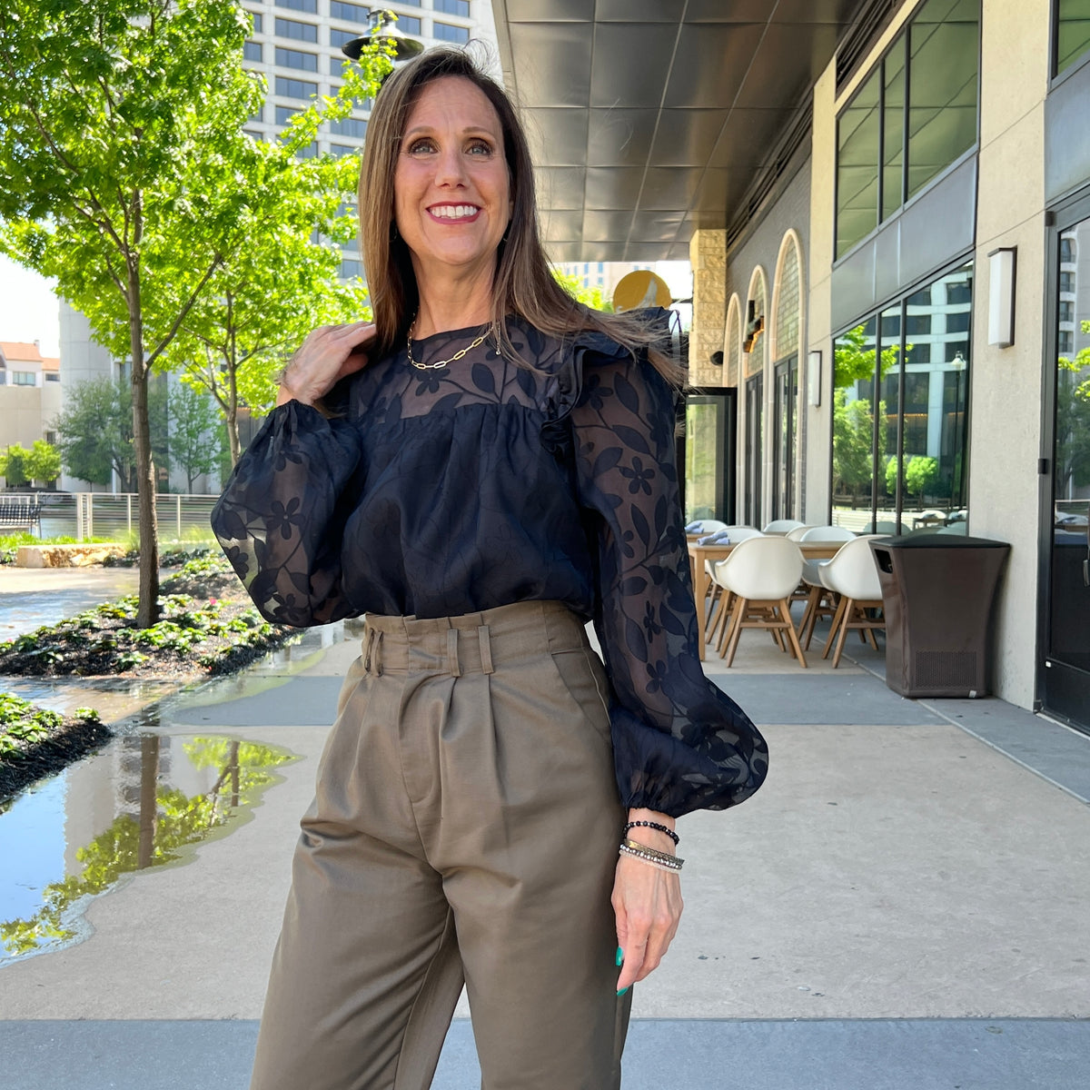 A woman standing in a plaza modeling a black shirt. The shirt has sheer black floral long sleeves and top with ruffles at the shoulders. She is wearing it tucked into a pair of olive pants.