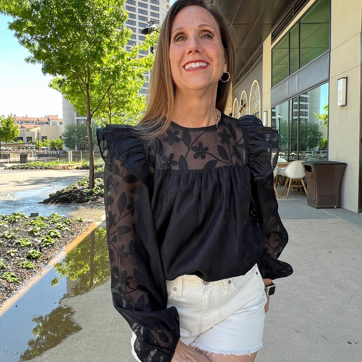 A woman standing in a plaza modeling a black shirt. The shirt has sheer black floral long sleeves and top with ruffles at the shoulders. She is wearing it tucked into a pair of white denim shorts.