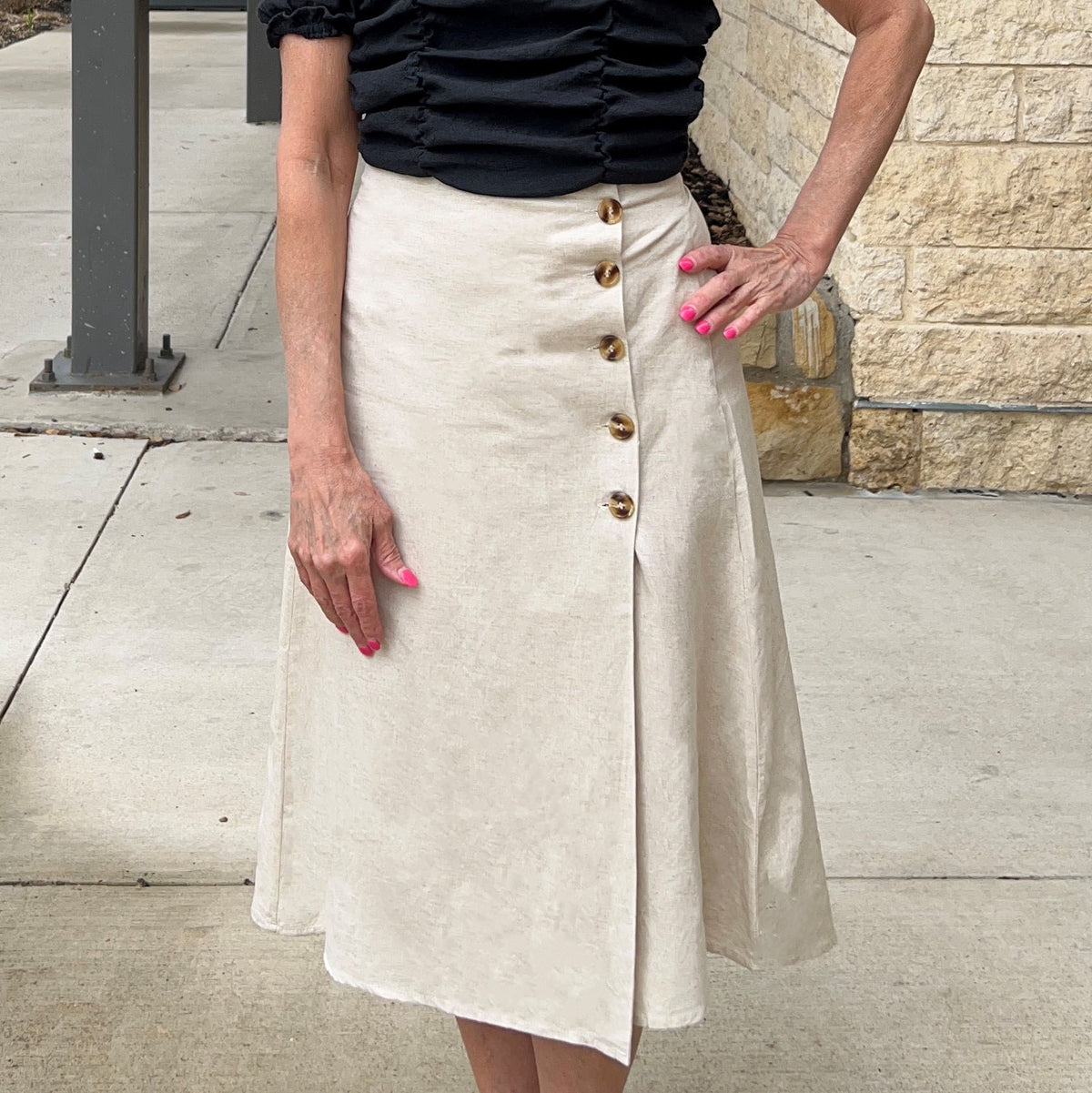 Beige oatmeal colored midi skirt with a wrap front. Tortoise shell buttons button the front side. Model is wearing it with a black crop top and is standing on a sidewalk. 