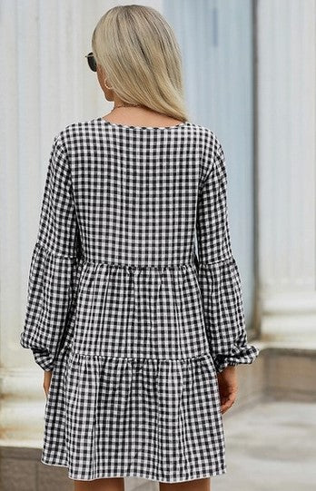 A woman standing on a sidewalk modeling the back of a three-tiered long-sleeved black and white gingham dress. 