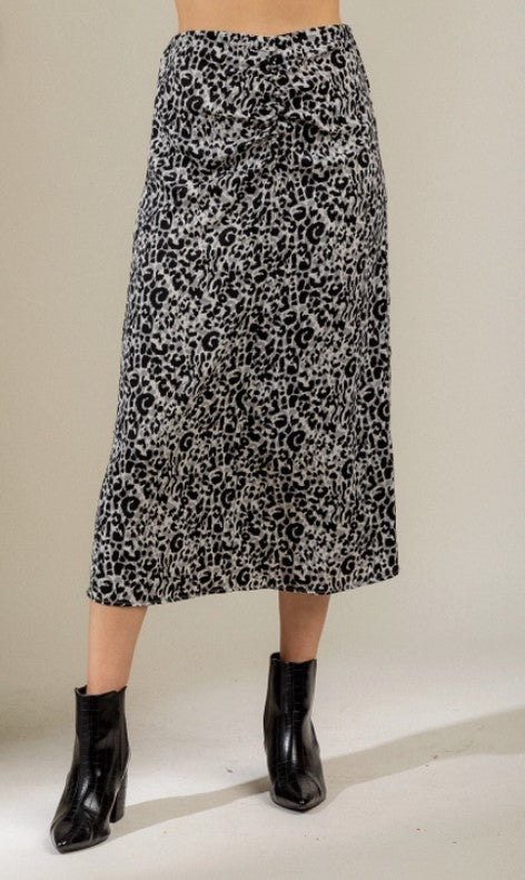 Midi length gray and black leopard print pencil skirt with a side zipper and ruched front. Model is standing in front of a beige  wall and is wearing black booties.