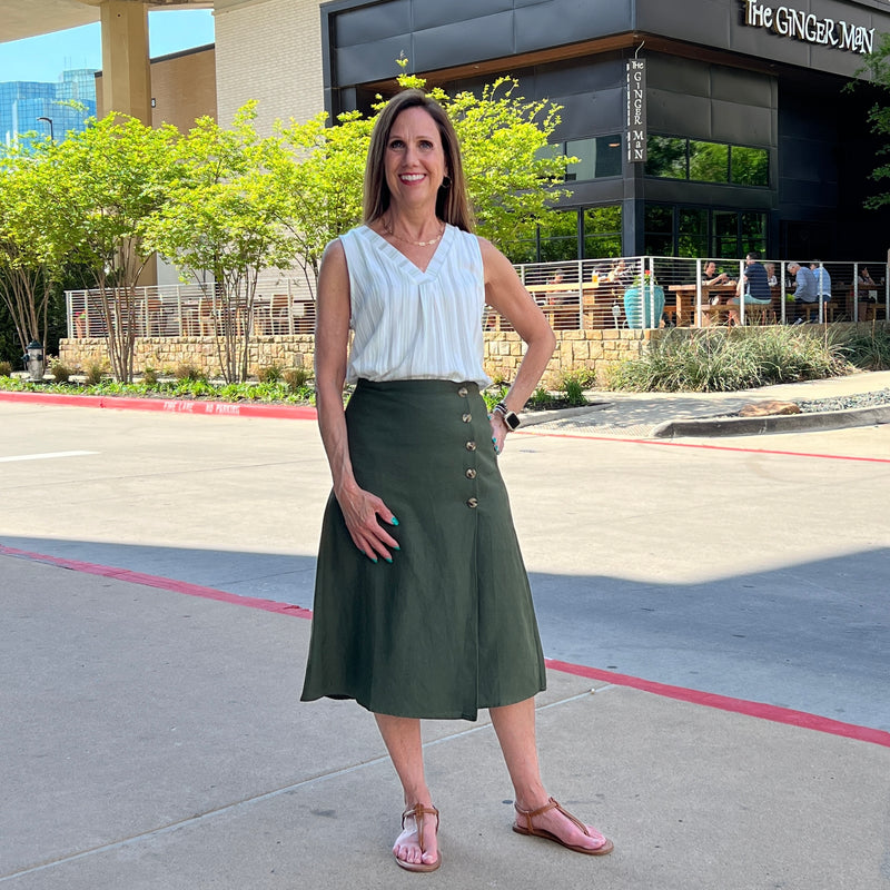 Forest olive green midi length skirt with a wrap front. Tortoise shell buttons button front on left side. Model is wearing a white blouse with olive stripes and is standing on a sidewalk. 