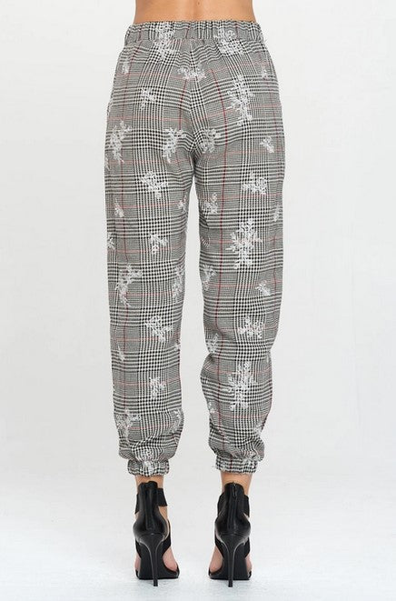 Woman modeling the back from the waist down of black, white and red plaid jogger pants with a white floral detail in front of a solid white background. She is also wearing black heeled sandals.