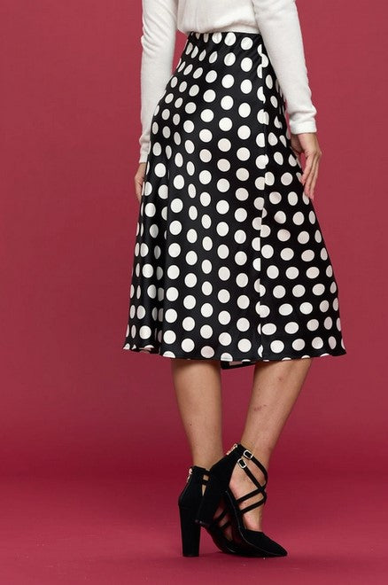Midi length black satin skirt with medium white polka dots. Pencil skirt shape at hips with a slight a-line flare at the knees to calves. Model is wearing the black satin skirt with black heels and white long-sleeve top. She standing in front of a red wall.